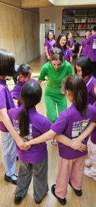 Kinder in violetten T-Shirts halten Händchen im Kreis, eine Frau in Grün leitet sie an.